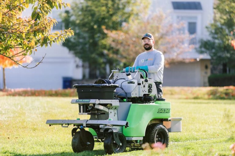 Lawn care technician riding a stand-on spreader across a residential lawn